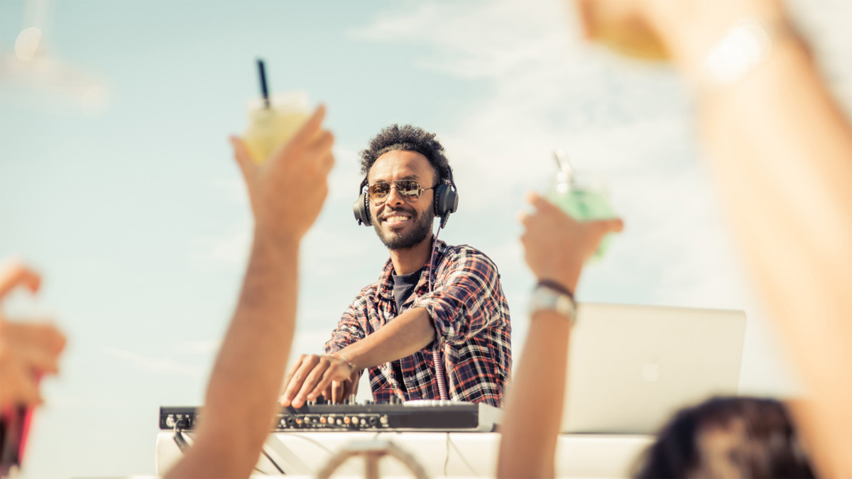 A DJ in sunglasses uses a soundboard while people dance with their hands in the air.