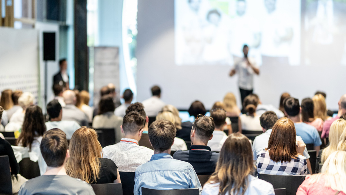 An audience of seated people observe as a man gives a presentation.