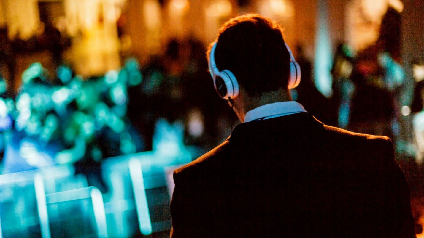 A wedding DJ wearing white headphones, wedding guests in the background.